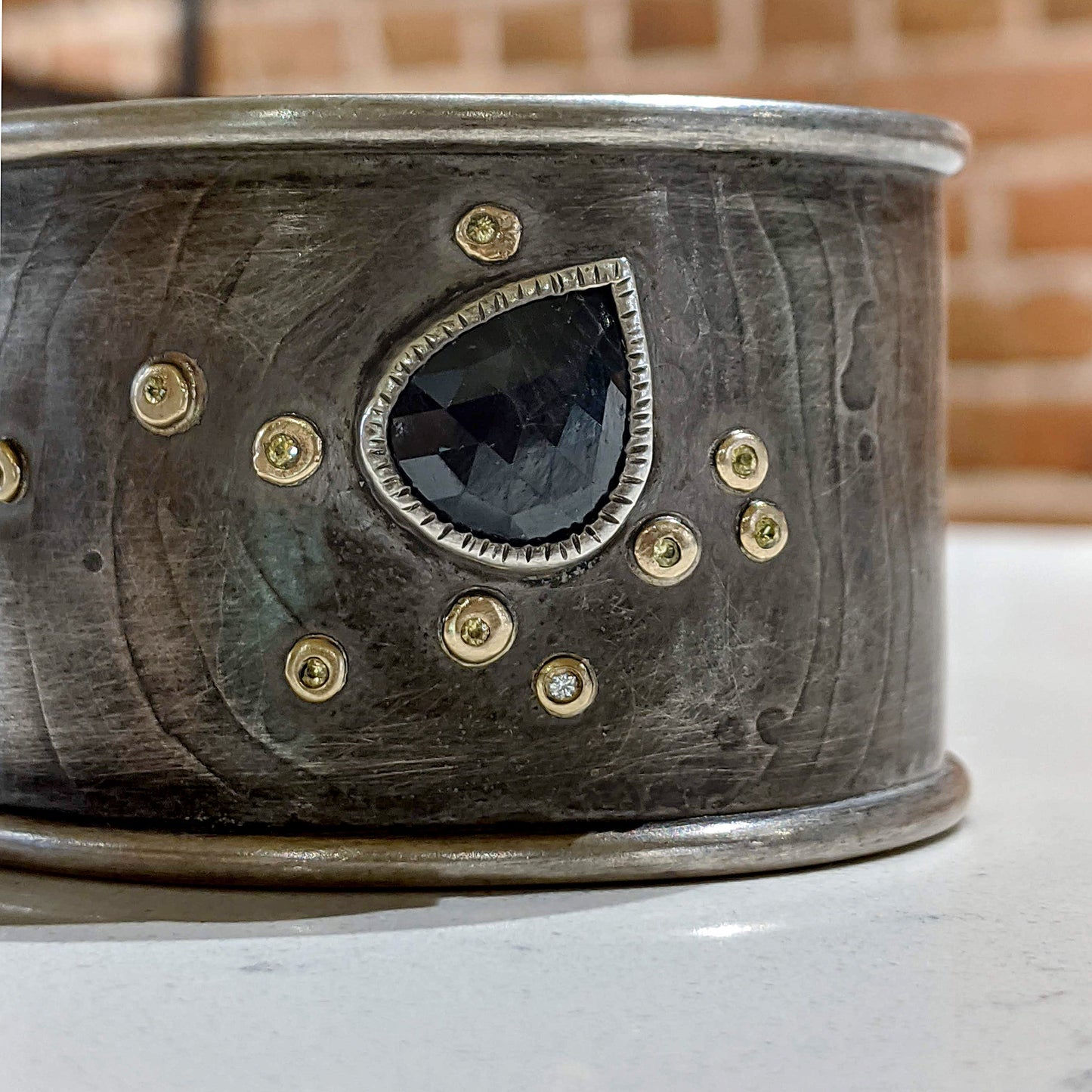 Cropped view of front of Sapphire Cuff Bracelet. Subtly Etched texture on a wide Sterling Cuff.  A Blue Rose Cut Sapphire is coupled with dots of 14K yellow gold surrounding small Yellow Sapphires and Diamonds.  