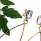 Detail photo of the top section of a pair of gold earrings a cluster of three white topaz gemstone set in an organic prong setting with a plant in the background.