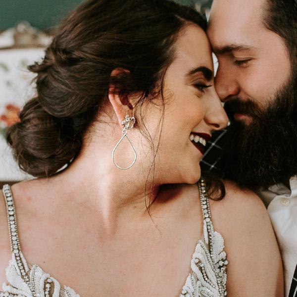 A woman and man smiling their foreheads touching in a lovingly manner. The woman is wearing a long and dramatic pair of earrings.