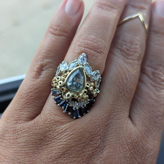 Close-up of a hand wearing an ornate ring with a large pear shaped natural gray diamond, sapphires, white diamonds and two tone white and yellow gold accents.
