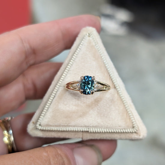 Custom Designed engagement Ring with a blue GREEN Montana Sapphire  and small diamonds in a triangle-shaped box held by a hand.