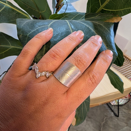 Hand wearing a silver ring with a diamond and a metallic leaf design, holding a green leafy plant.