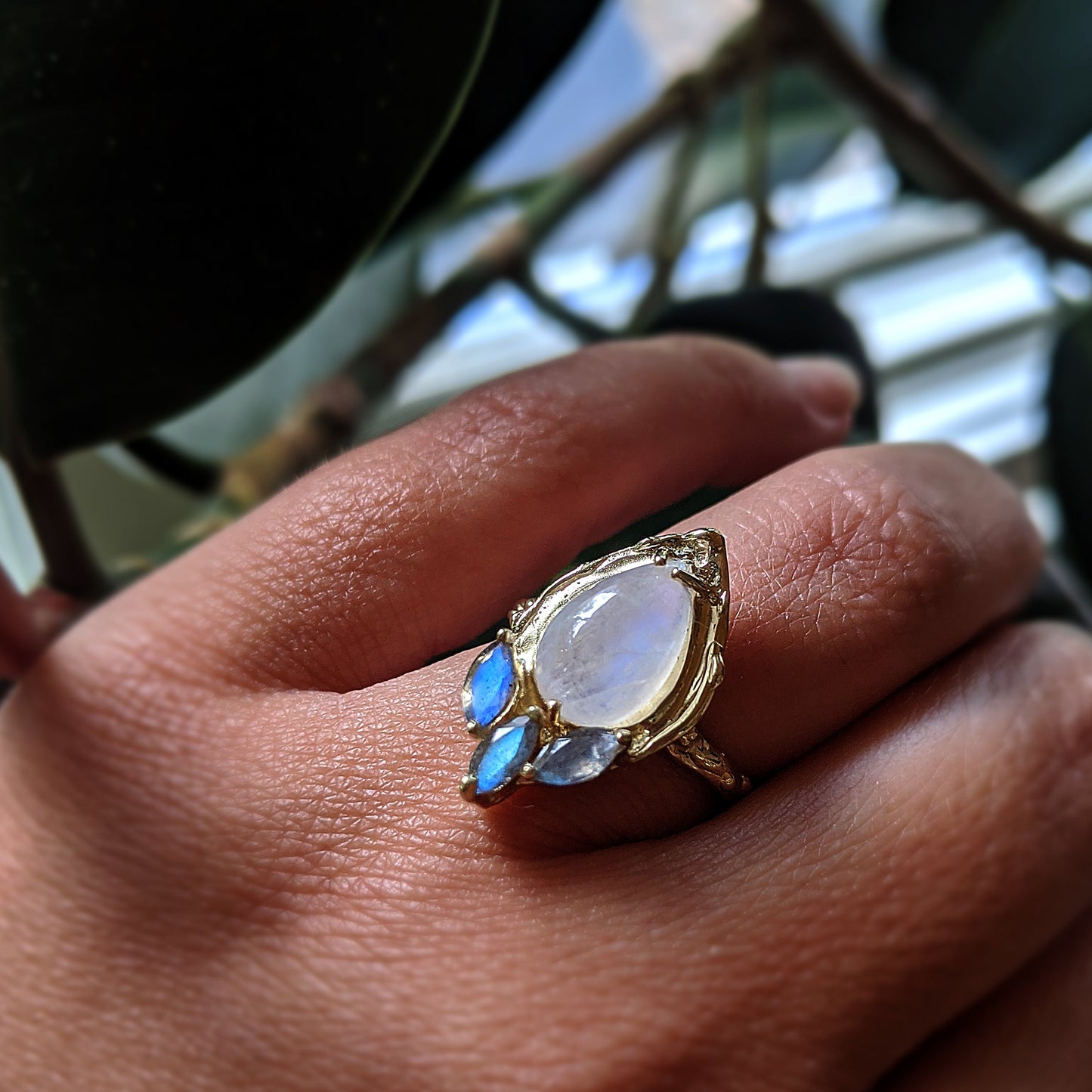 Full view of Celeste Ring on woman's hand to help give an idea of its scale. This ring is made of yellow gold and has a set teardrop shaped moonstone and three smaller set laboradites underneath the arch of the moonstone.