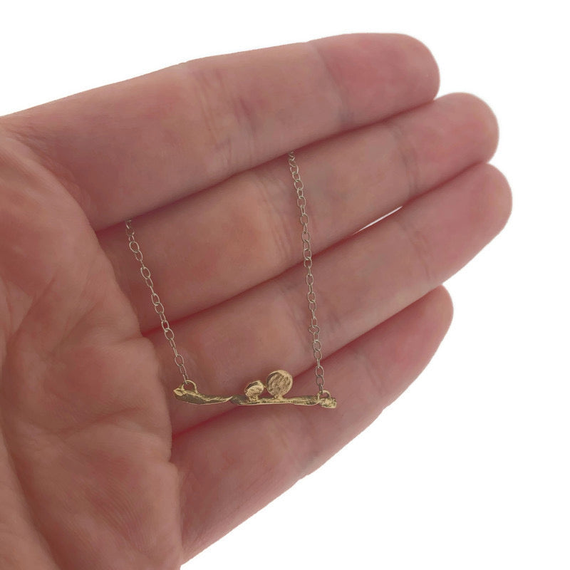 Close up view of pendant on Blank Bar Necklace with hand in background to help give idea of scale of piece. This pendant is made in yellow gold and lays on a silver chain. The pendant is in the shape of a horizontal line that resembles a branch that has two dots of gold on top of the branch.