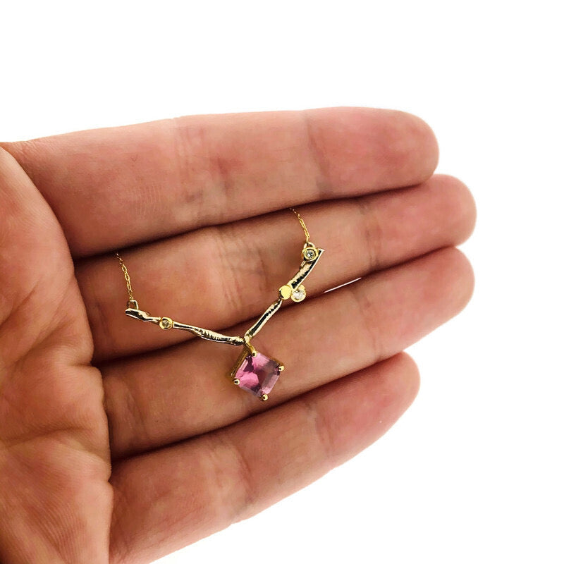 Close up image of Amelie Necklace with hand in background to help give idea of scale of piece. This piece is made of gold and lays on a gold chain. The pendant is in the shape of a "v" and has characteristics of a branch with three diamonds set on its outer corner. At the tip of the "v" is a set square pink sapphire.