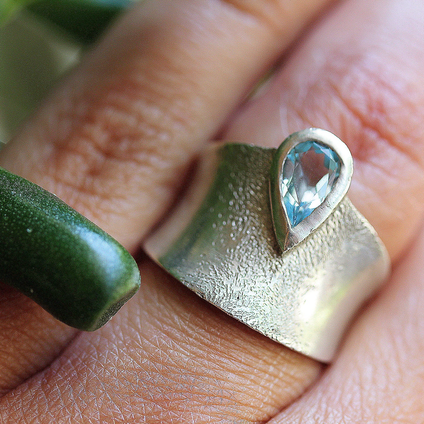 Full view of Astoria Ring on woman's hand to help give an idea of its scale. A bezel set blue Topaz is perched slightly off center along the edge of a gradually sweeping concave band, surrounded by a cluster of dappled texture. 