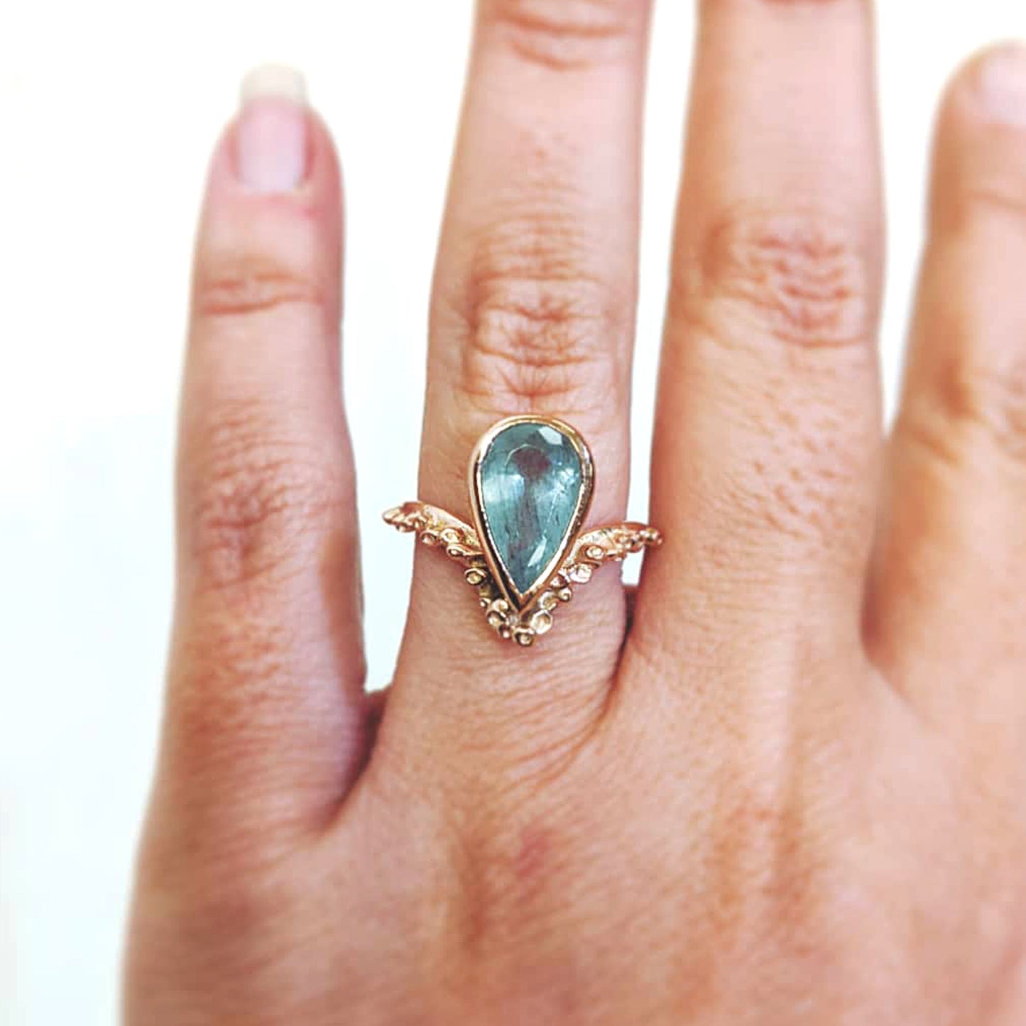 Full view of Piper Ring. This ring is made of gold, features a teardrop shaped moss aquamarine bezel set with Katie's signature seasponge texture surrounding the tip of the teardrop and continuing on the band.