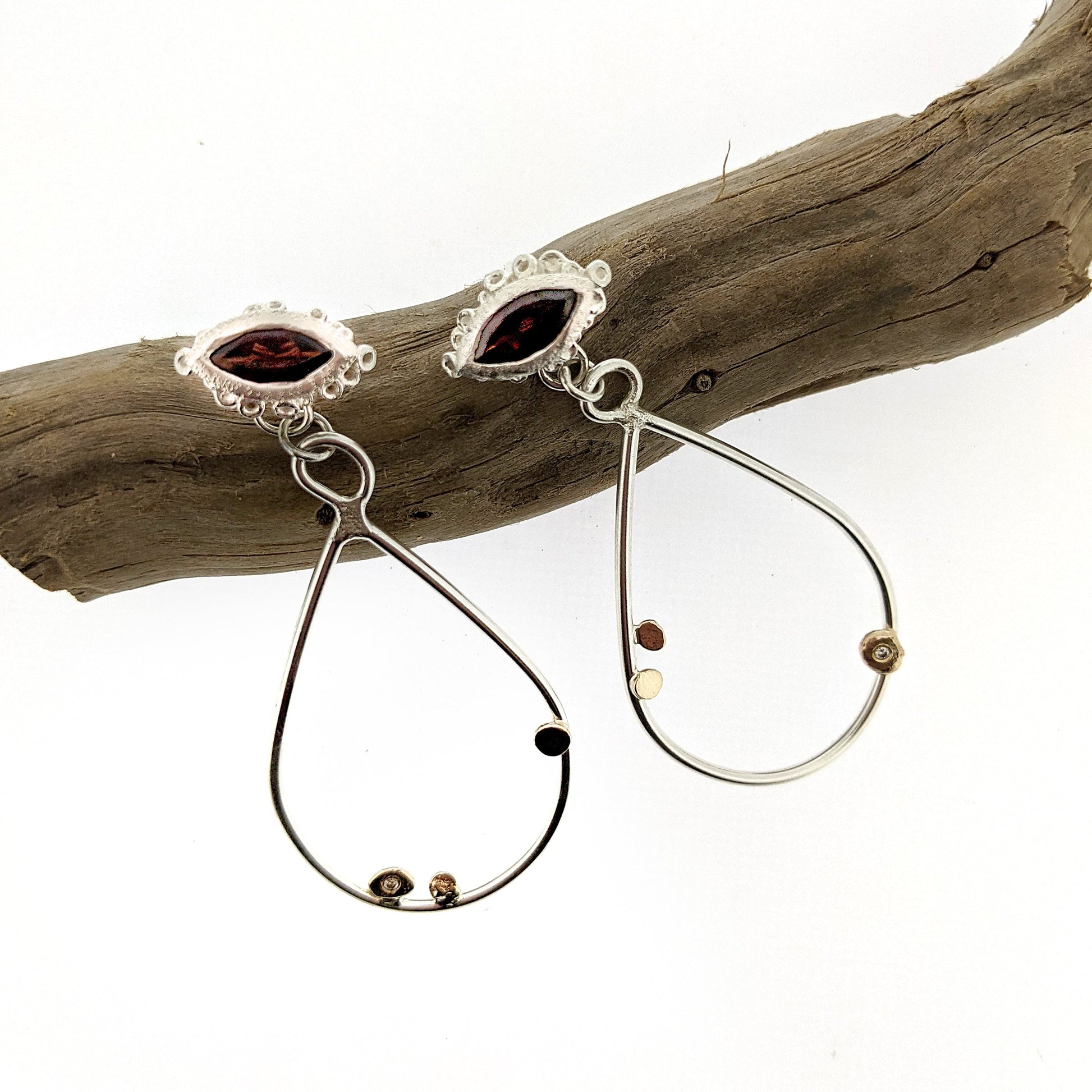 Full view of Trena Earring on stick. These dangle earrings have a marquise cut garnet surrounded by Katie's signature seasponge texture with a teardrop of silver wire hanging off them that has a few gold dots and set diamonds on it.