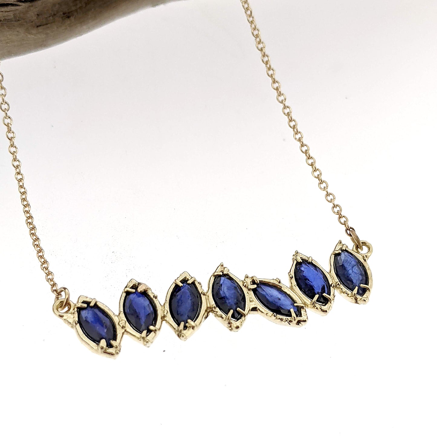 Close up view of pendant on Sapphire Cherin Necklace. Seven Sparkling natural marquise shaped Blue Sapphire stones delicately adorn this irregular line-style bar necklace with subtle organic detailing. 
