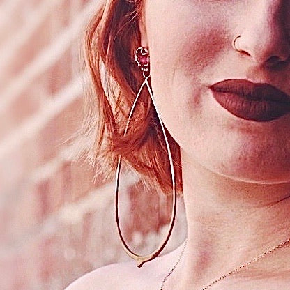 Full view of Louisa Earrings being worn to help give an idea of their scale. They are made of sterling silver, dipped in gold powdercoat, have a set ruby at the top and dangling from that a teardrop shaped piece of wire.