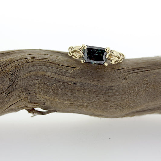 Full view of Kendra Moissanite Ring laying on top of stick. One of a kind Organic modern engagement ring featuring a large 1.88 Carat dark blue green radiant cut Moissanite set in 14K yellow gold.      As unique a solitaire as you'll find, with delicate botanical details on each side, framing the center stone.  