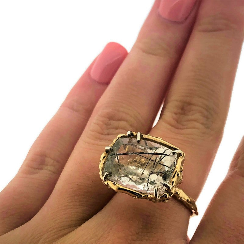Full view of Tourmilated Quartz Organic Ring on woman's hand to help give an idea of its scale. This gold ring showcases a rectangle shaped tourmilated quarts set with seven silver prongs on top of a gold basket and band that were both hand carved with an organic nature.